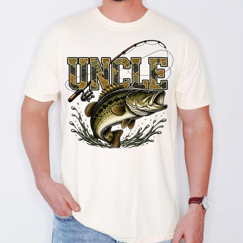 K&ouml;nnte beinhalten: Cremefarbenes T-Shirt mit einer Grafik eines Gro&szlig;maulbasses, der aus dem Wasser springt, einer Angelrute und dem Wort "UNCLE" in Tarnschrift. Das Design ist mittig auf dem Shirt platziert.