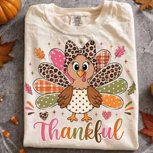 May include: A cream-colored t-shirt featuring a cartoon turkey graphic with a leopard print bow. The turkey has multicolored feathers with polka dot, plaid, and leopard patterns. The word Thankful is printed in a colorful script font below.