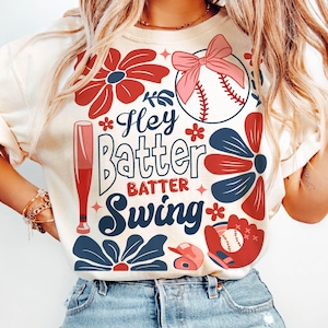 May include: A white t-shirt with a baseball-themed design. The design features a baseball, bat, glove, and flowers in red, blue, and pink. The text on the shirt reads "Hey Batter Batter Swing".