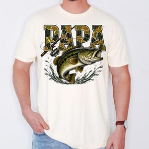 K&ouml;nnte beinhalten: Cremefarbenes T-Shirt mit dem Wort "PAPA" in Tarnschrift &uuml;ber einer Grafik eines Gro&szlig;maulbasses, der aus dem Wasser springt. Eine Angelrute ist ebenfalls abgebildet. Das Shirt ist ein Freizeitkleidungsst&uuml;ck.