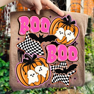Puede incluir: Una camiseta marr&oacute;n con un dise&ntilde;o de Halloween. El dise&ntilde;o incluye la palabra "BOO" en letras rosas, calabazas naranjas, fantasmas blancos, murci&eacute;lagos negros y estrellas a cuadros. El tema general es espeluznante y divertido.