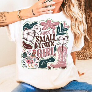 Pu&ograve; includere: T-shirt bianco con un design grafico colorato. Il design include il testo "Just a Small Town Girl" con uno stivale da cowboy, fiori di cotone, un gallo ed elementi floreali in rosa, verde e marrone.