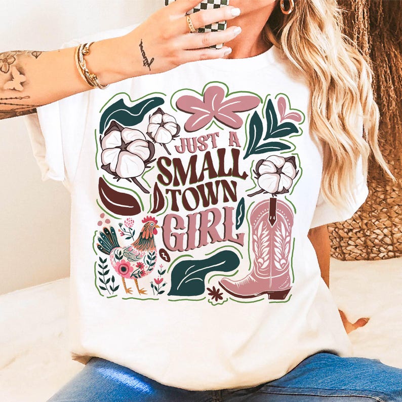 Pu&ograve; includere: T-shirt bianco con un design grafico colorato. Il design include il testo "Just a Small Town Girl" con uno stivale da cowboy, fiori di cotone, un gallo ed elementi floreali in rosa, verde e marrone.