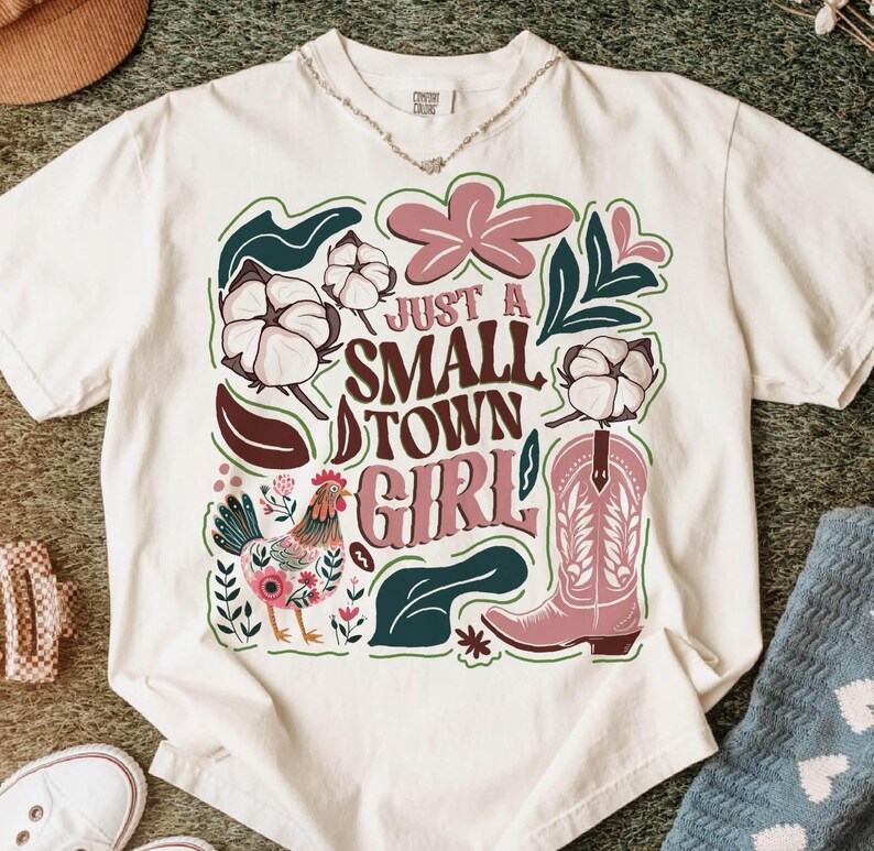 Pu&ograve; includere: T-shirt bianco con un design grafico colorato. Il design include il testo "Just a Small Town Girl" con illustrazioni di cotone, uno stivale da cowboy rosa, un gallo ed elementi floreali. La maglietta ha uno scollo tondo.
