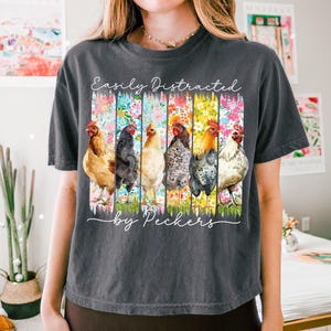 May include: Dark gray t-shirt featuring a watercolor design of six chickens, each in a different color, set against vertical floral stripes. The text "Easily Distracted by Peckers" is written in a script font.