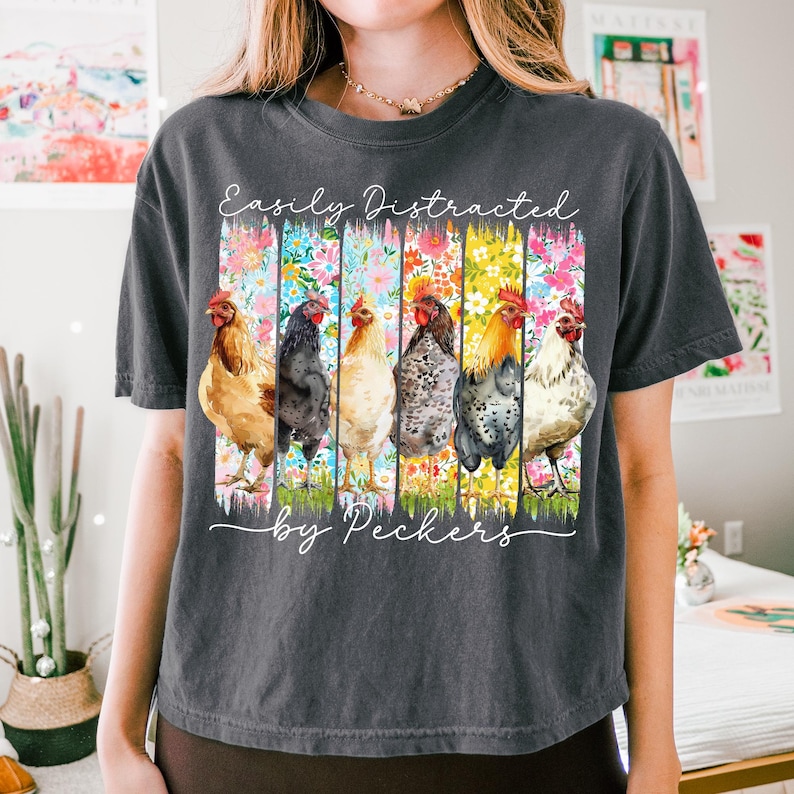 May include: Dark gray t-shirt featuring a watercolor design of six chickens, each in a different color, set against vertical floral stripes. The text "Easily Distracted by Peckers" is written in a script font.