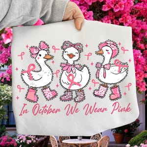 Può includere: Un oggetto di tessuto bianco con tre anatre dei cartoni animati con fiocchi, stivali e nastri rosa, con la scritta "In October We Wear Pink". Le anatre camminano, circondate da nastri rosa.