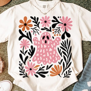 May include: White t-shirt featuring a pink ghost design surrounded by orange, pink, and black floral elements. The ghost has a dotted pattern and a simple face. The shirt has a crew neck and short sleeves.