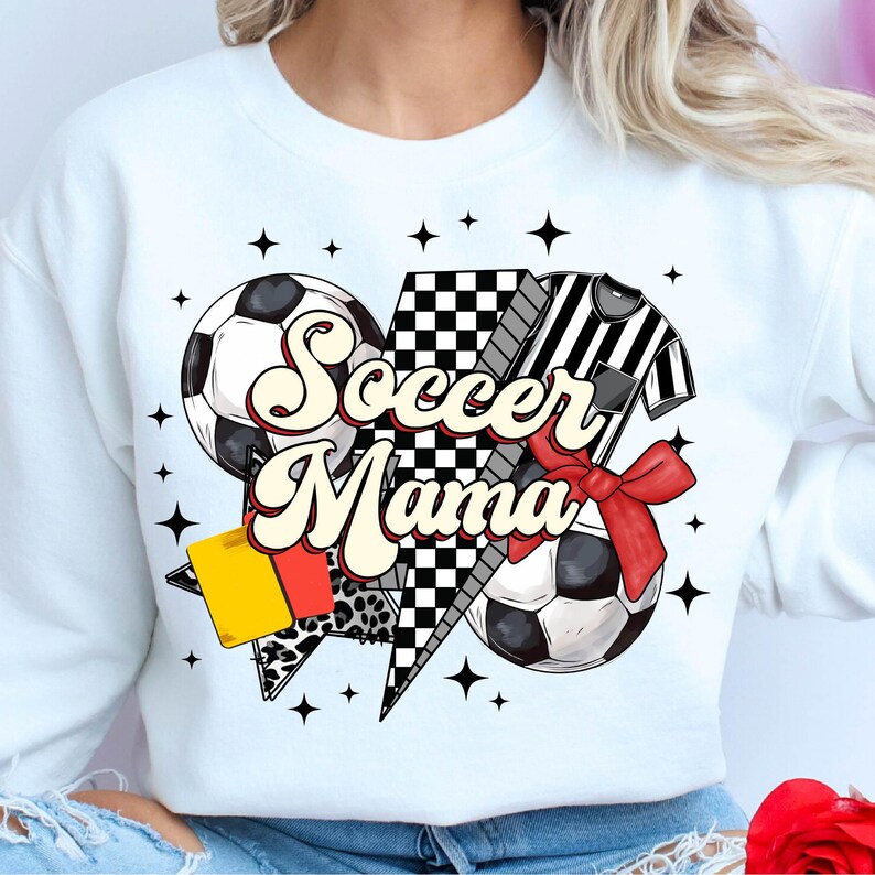 K&ouml;nnte beinhalten: Wei&szlig;es Sweatshirt mit einem Grafikdesign aus einem Fu&szlig;ball, einem Schiedsrichtertrikot und einer karierten Flagge. Der Text "Soccer Mama" ist in einer Retro-Schrift geschrieben.