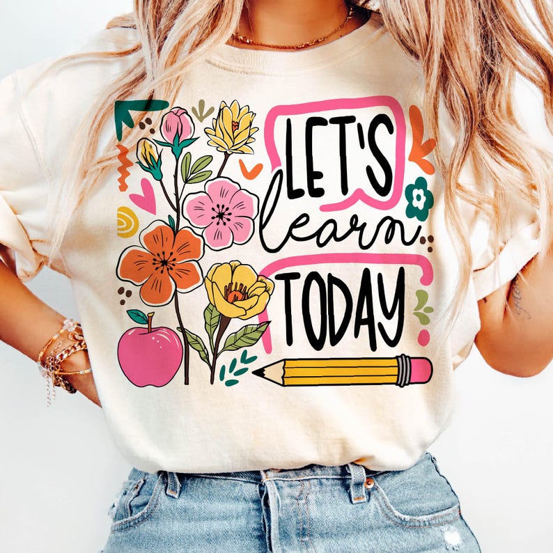 May include: Cream-colored sweatshirt with a colorful graphic. The design includes the text "LET'S learn TODAY" in black, surrounded by a pink border. Floral and apple illustrations are also present, along with a yellow pencil.
