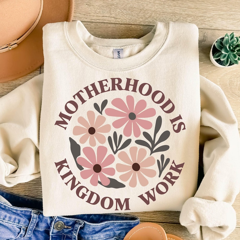 Puede incluir: Una sudadera de color crema con el texto "Motherhood is Kingdom Work" en letras marrones. El texto est&aacute; rodeado por un c&iacute;rculo de flores rosas y hojas grises.