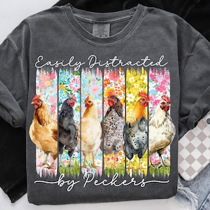 May include: Dark gray t-shirt with the text "Easily Distracted by Peckers." The shirt features a colorful floral design with six different chickens. The chickens are in various colors, including brown, black, and white.