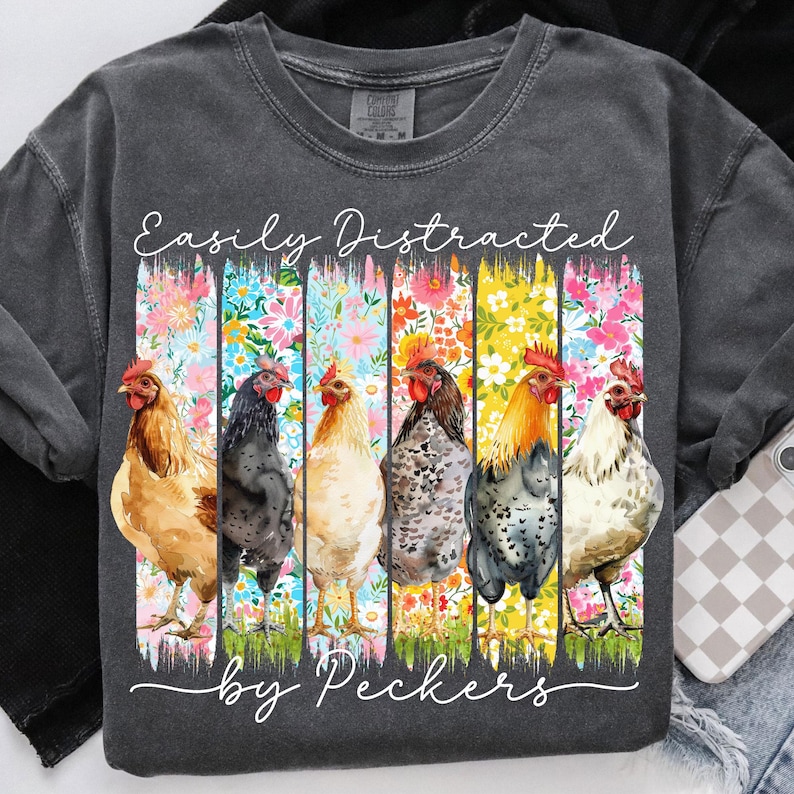 May include: Dark gray t-shirt with the text "Easily Distracted by Peckers." The shirt features a colorful floral design with six different chickens. The chickens are in various colors, including brown, black, and white.