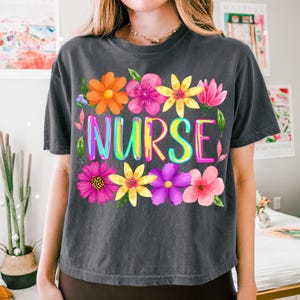 以下が含まれることがあります： カラフルな文字で「NURSE」と書かれた、鮮やかな花柄のダークグレーのTシャツ。オレンジ、ピンク、黄色、紫の色合いの花が描かれています。Tシャツはクルーネックで半袖です。