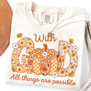 May include: Cream-colored t-shirt with the words "With God All things are possible" in orange script. The letters "G" and "D" are filled with floral patterns. A pumpkin with a bow and daisies is in the center.