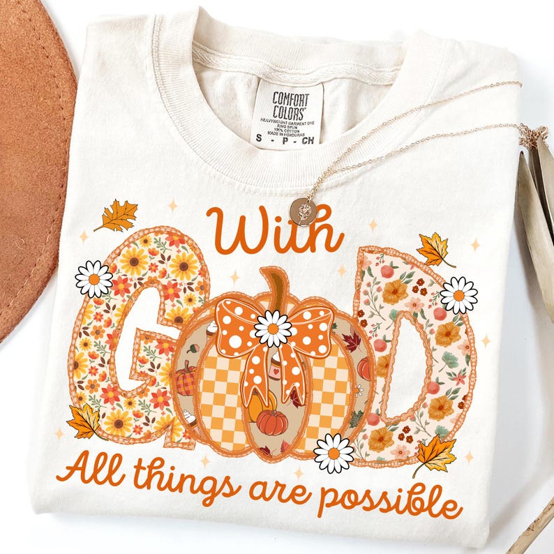 May include: Cream-colored t-shirt with the words "With God All things are possible" in orange script. The letters "G" and "D" are filled with floral patterns. A pumpkin with a bow and daisies is in the center.