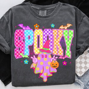May include: Dark gray long-sleeve shirt with a colorful "SPOOKY" graphic. The graphic features a ghost character with a witch hat, holding a jack-o'-lantern, and surrounded by bats and flowers. The word "SPOOKY" is in a checkered pattern.