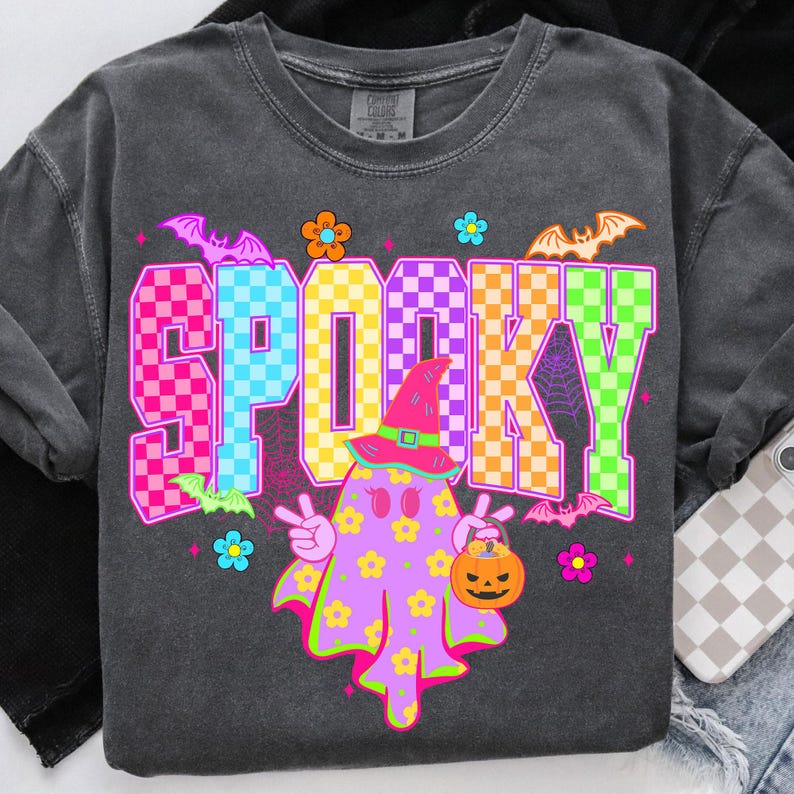 May include: Dark gray long-sleeve shirt with a colorful "SPOOKY" graphic. The graphic features a ghost character with a witch hat, holding a jack-o'-lantern, and surrounded by bats and flowers. The word "SPOOKY" is in a checkered pattern.