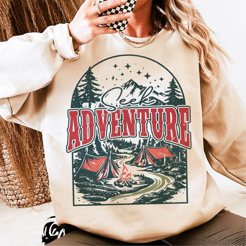 May include: A beige sweatshirt with a vintage-style graphic print of a mountain scene with a campfire, trees, and the text "Seek Adventure".