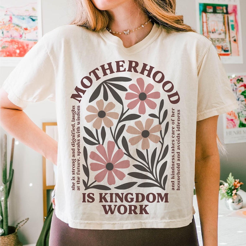 Puede incluir: Una camiseta blanca con un dise&ntilde;o gr&aacute;fico marr&oacute;n que dice "MOTHERHOOD IS KINGDOM WORK" con un dise&ntilde;o floral y un texto que dice "Ella es fuerte y digna, se r&iacute;e del futuro, habla con sabidur&iacute;a y amabilidad, cuida de su hogar y evita la pereza."
