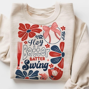May include: A beige sweatshirt with a baseball-themed design. The design features a baseball, a bat, flowers, and the text "Hey Batter Batter Swing".