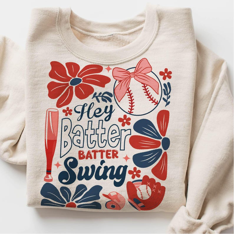 May include: A beige sweatshirt with a baseball-themed design. The design features a baseball, a bat, flowers, and the text "Hey Batter Batter Swing".
