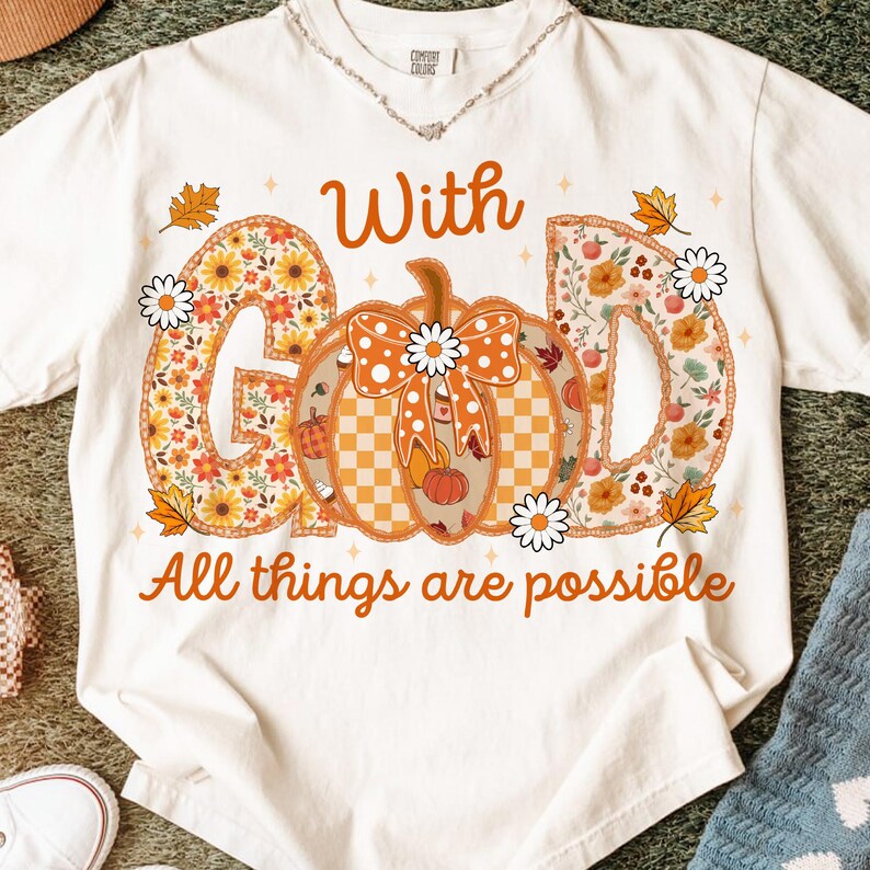 May include: White t-shirt with the words "With God All things are possible" in orange. The word "God" is designed with floral patterns and a pumpkin in the center. Daisies and fall leaves decorate the design.