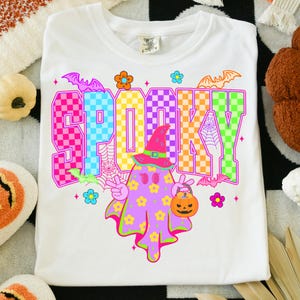 May include: White t-shirt with the word "SPOOKY" in colorful checkered letters. A cartoon ghost with a witch hat and a pumpkin bucket is in the center. Bats and flowers surround the design.