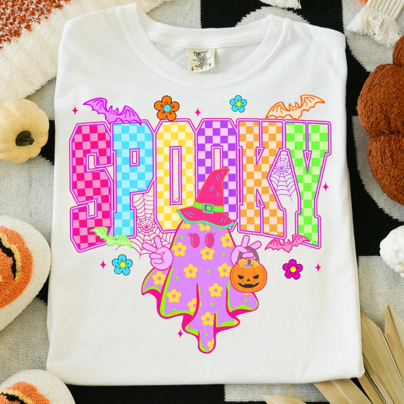 May include: White t-shirt with the word "SPOOKY" in colorful checkered letters. A cartoon ghost with a witch hat and a pumpkin bucket is in the center. Bats and flowers surround the design.