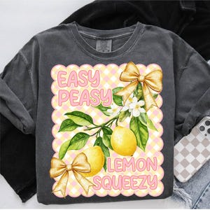 Pu&ograve; includere: Una maglietta grigia con un design grafico rosa e giallo. Il grafico presenta limoni, fiori e fiocchi. Il testo "Easy Peasy Lemon Squeezy" &egrave; scritto in lettere rosa.