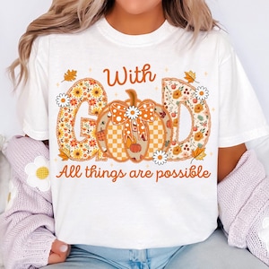 May include: White t-shirt with the text "With God All things are possible." The word "GOD" is designed with a floral pattern. A pumpkin with a bow is in the center. The shirt has a fall theme.