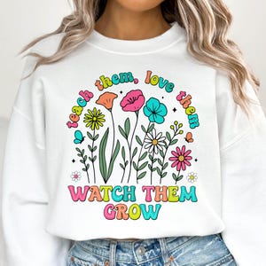 Puede incluir: Sudadera blanca con un dise&ntilde;o gr&aacute;fico colorido con flores y el texto "Teach them, love them, watch them grow".