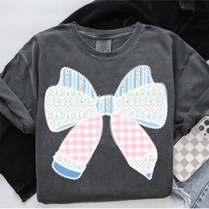May include: A grey t-shirt featuring a large, blue, pink and white bow design. The bow is made from various patterned fabrics, including gingham, floral and stripes.