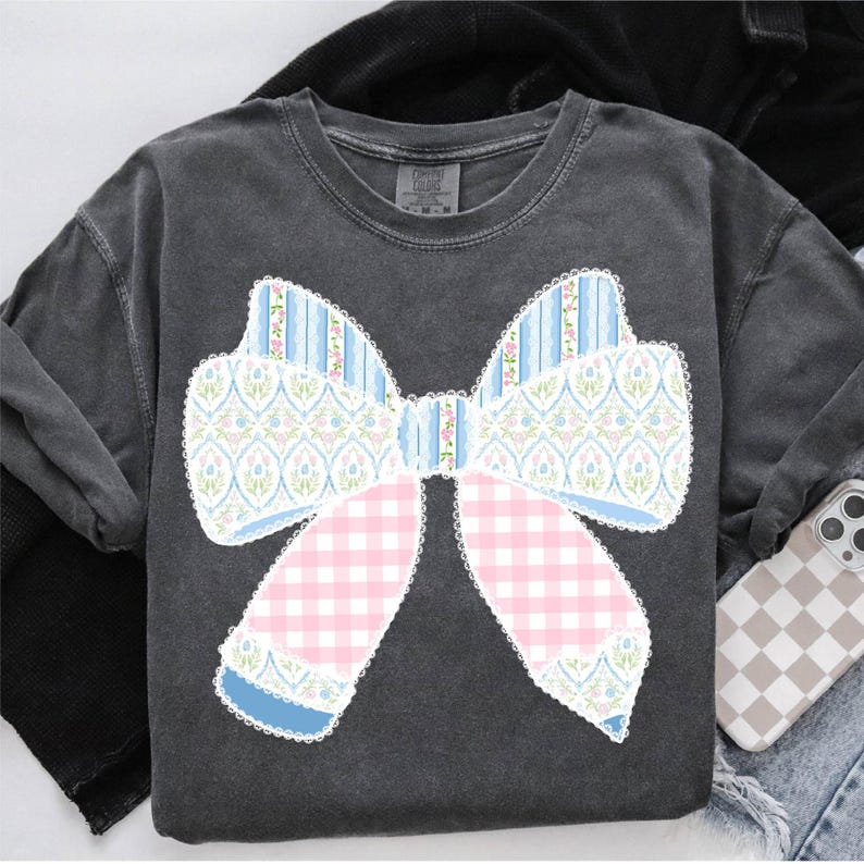 May include: A grey t-shirt featuring a large, blue, pink and white bow design. The bow is made from various patterned fabrics, including gingham, floral and stripes.