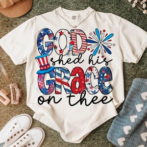 May include: Cream-colored t-shirt with the phrase "GOD shed his GRACE on thee" in patriotic red, white, and blue lettering. The design incorporates stars, stripes, and a top hat, evoking a Fourth of July theme.