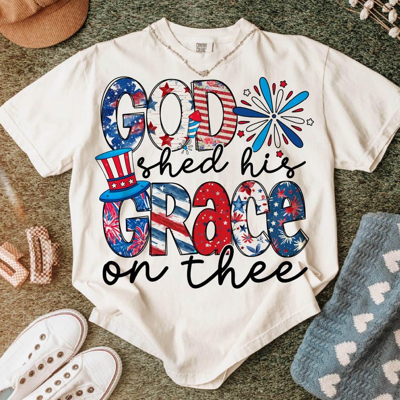 May include: Cream-colored t-shirt with the phrase "GOD shed his GRACE on thee" in patriotic red, white, and blue lettering. The design incorporates stars, stripes, and a top hat, evoking a Fourth of July theme.