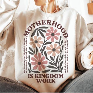 Puede incluir: Una sudadera blanca con un dise&ntilde;o floral y el texto "MOTHERHOOD IS KINGDOM WORK" impreso en la parte delantera. El texto est&aacute; en marr&oacute;n y negro, y las flores son rosas y marrones.