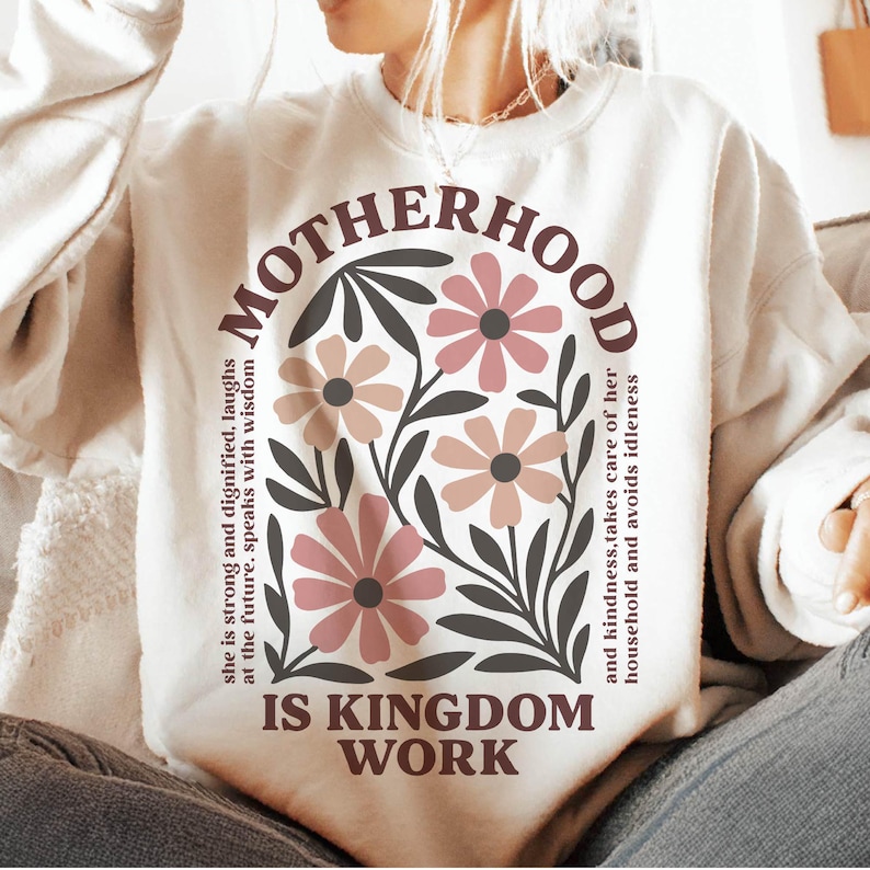 Puede incluir: Una sudadera blanca con un dise&ntilde;o floral y el texto "MOTHERHOOD IS KINGDOM WORK" impreso en la parte delantera. El texto est&aacute; en marr&oacute;n y negro, y las flores son rosas y marrones.