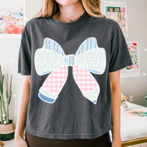 May include: A dark grey cropped t-shirt featuring a large, colourful bow design. The bow is made from various patterned fabrics, including pink and blue gingham, floral print, and striped fabric.