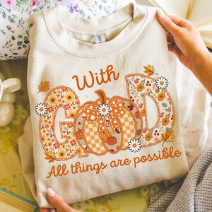 May include: Cream-colored sweatshirt with the words "With God" and "All things are possible" printed in orange. The design includes a pumpkin and floral letters, with fall leaves and daisy accents.