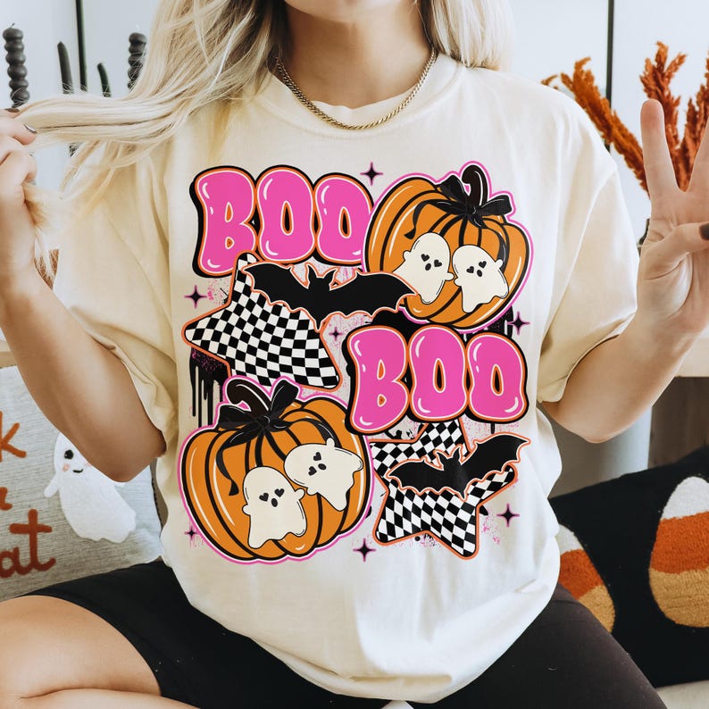 Puede incluir: Camiseta color crema con un dise&ntilde;o tem&aacute;tico de Halloween. El dise&ntilde;o presenta la palabra "BOO" en letras rosas con burbujas, calabazas, fantasmas, murci&eacute;lagos y estrellas a cuadros. La camiseta tiene un corte holgado.
