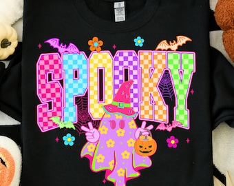 May include: Black sweatshirt with the word "SPOOKY" in colorful checkered letters. A ghost character wearing a witch hat and holding a jack-o'-lantern is featured below the word. Bats and flowers decorate the design, perfect for Halloween.