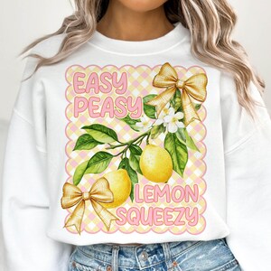 Pu&ograve; includere: Felpa bianca con un design a scacchi rosa e giallo. Il design presenta un albero di limone con limoni, fiori e fiocchi. Il testo "Easy Peasy Lemon Squeezy" &egrave; scritto in rosa.