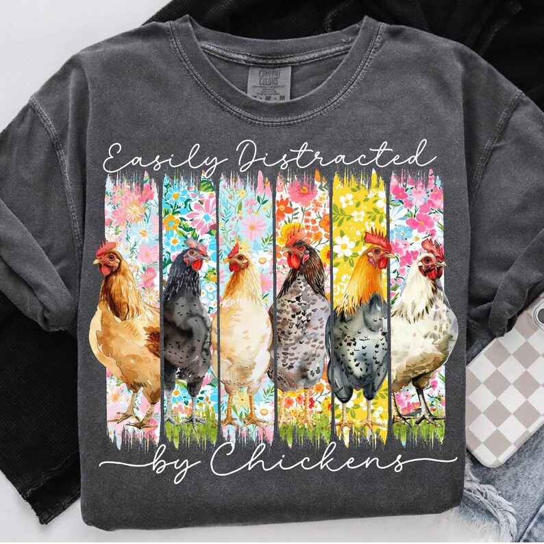 以下が含まれることがあります： カラフルな花柄と「Easily Distracted by Chickens」の文字が入った濃いグレーのスウェットシャツ。 グラフィックには、それぞれユニークな羽の模様と色を持つ6羽の水彩画の鶏が描かれています。