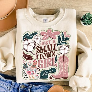 Pu&ograve; includere: Felpa color crema con un design grafico. Il design include il testo "Just a Small Town Girl", uno stivale da cowboy rosa, fiori di cotone, un gallo ed elementi floreali rosa e verdi.