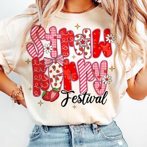 May include: A white t-shirt with a red and pink graphic design that says "Strawberry Festival". The design features strawberries, stars, and flowers.