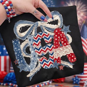 Könnte beinhalten: Schwarzes quadratisches Kissen mit den Buchstaben "USA" aus rot, weiß und blauem Stoff. Die Buchstaben sind mit Sternen, Spitze und einer großen Schleife verziert. Patriotisches Thema.