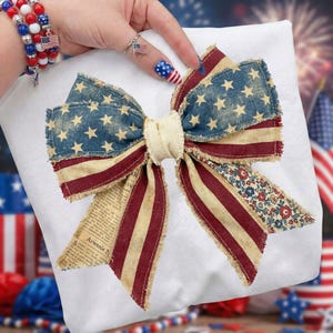 May include: A patriotic bow featuring the American flag design. The bow has a blue section with white stars, red and white stripes, and a floral print. The bow is made of fabric and has a frayed edge.