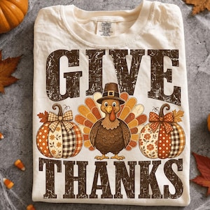 Op de afbeelding: Een crèmekleurig t-shirt met de tekst GIVE THANKS in grote, verweerde bruine letters. Tussen de woorden staat een centrale illustratie van een kalkoen met een pelgrimshoed, geflankeerd door twee pompoenen met ruit- en bloemenpatronen.