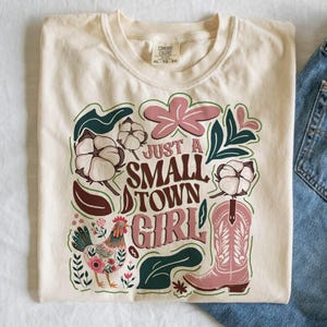 Pu&ograve; includere: T-shirt color crema con la scritta "Just a Small Town Girl" in un carattere decorativo. Il design include uno stivale da cowboy rosa, fiori di cotone, un gallo e accenti floreali. La maglietta &egrave; piegata.